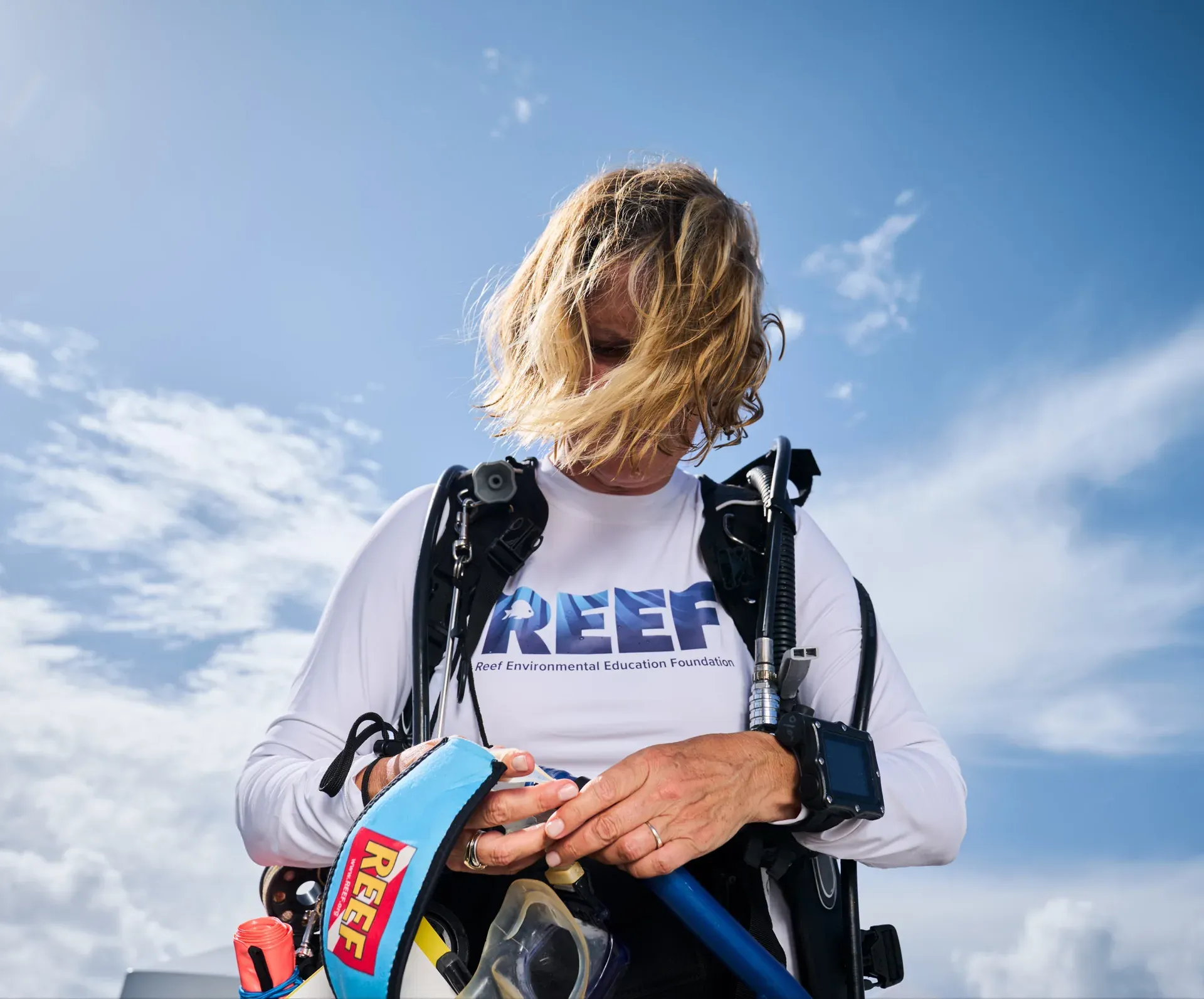 A diver in a REEF Environmental Education Foundation rash guard checking dive gear against a bright blue sky